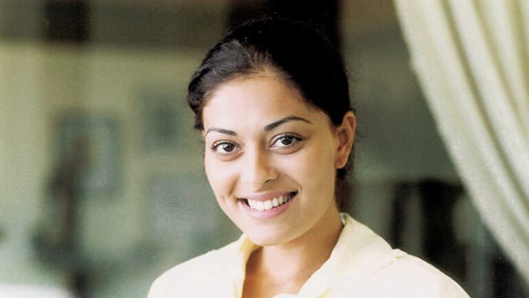 Mulher jovem sorrindo em um retrato iluminado de forma suave. Ela tem cabelos escuros presos para trás, covinhas discretas e veste uma camisa clara. O fundo desfocado sugere um ambiente interno, transmitindo leveza e naturalidade.