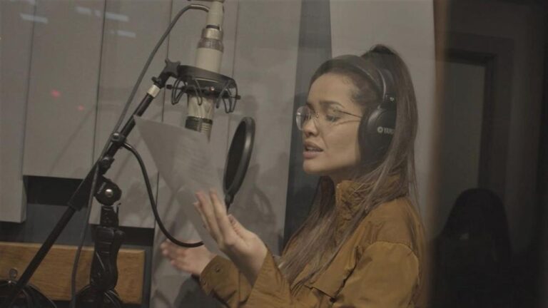Mulher em estúdio de gravação, usando fones de ouvido grandes e óculos, canta ou narra enquanto segura uma folha de papel diante de um microfone profissional suspenso. O ambiente é iluminado de forma suave, com paredes acústicas ao fundo.