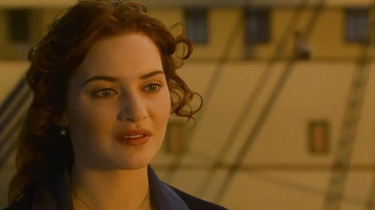 Close-up de uma mulher de cabelos ruivos ondulados, em cena a bordo de um navio, iluminada por luz suave e com expressão contemplativa.