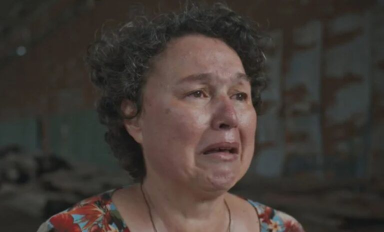 Mulher de meia-idade com cabelos curtos e cacheados aparece chorando, com expressão de dor e sofrimento. Seu rosto está avermelhado e molhado por lágrimas. Ela veste uma blusa estampada com flores e está em um ambiente interno escuro e desgastado ao fundo
