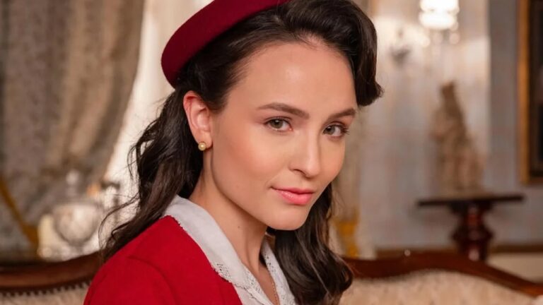 Jovem mulher com expressão serena posa em ambiente elegante. Ela usa boina vermelha, vestido vermelho com gola branca rendada e brincos de pérola. O fundo desfocado revela móveis clássicos e iluminação suave, sugerindo cenário de época.