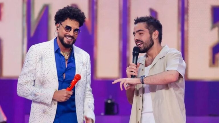 Dois homens estão em um palco colorido com iluminação roxa e microfones nas mãos — um veste paletó branco com brilhos, camisa azul e óculos escuros, enquanto o outro usa camisa bege clara sobre camiseta branca, conversando e sorrindo durante uma apresentação.