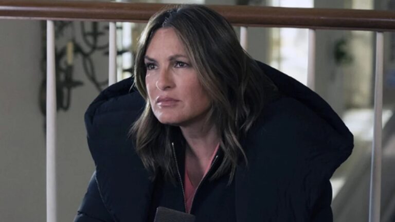 Mulher de expressão séria e olhar atento usa casaco grosso preto e blusa rosa por baixo, com cabelos castanhos soltos na altura dos ombros, sentada em ambiente interno iluminado com corrimão branco e fundo desfocado