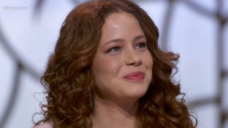 Mulher jovem com cabelos ruivos cacheados sorri levemente em um estúdio iluminado, olhando para o lado; ela usa maquiagem suave e brincos delicados, com o fundo desfocado exibindo formas geométricas claras.