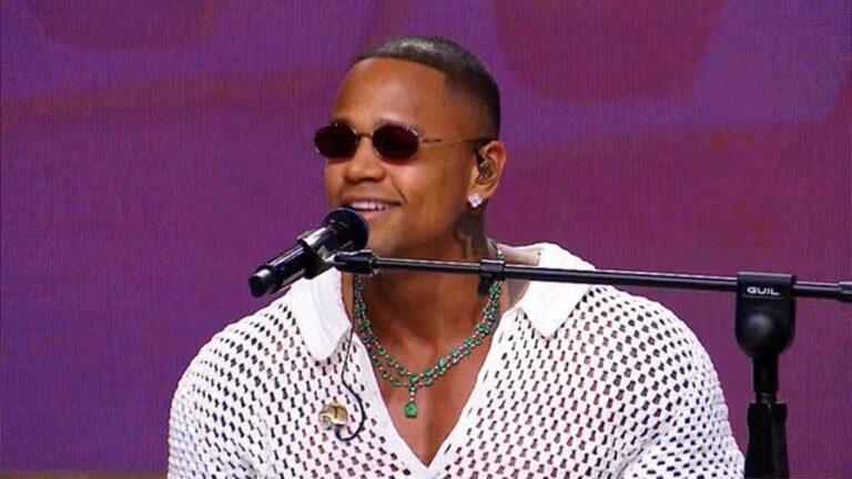 Homem usa óculos escuros, colar verde e camisa branca vazada enquanto canta ou fala ao microfone em estúdio com fundo roxo