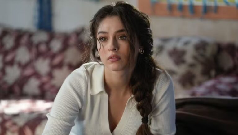 Mulher de expressão pensativa e olhar fixo à frente usa blusa branca de manga longa e cabelo preso em trança lateral solta, sentada em ambiente interno com sofá estampado e luz natural entrando pela janela ao fundo.