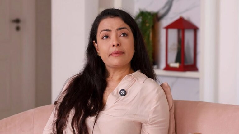 Mulher sentada em sofá cor-de-rosa com expressão emocionada em ambiente iluminado decorado com plantas e lanterna vermelha ao fundo