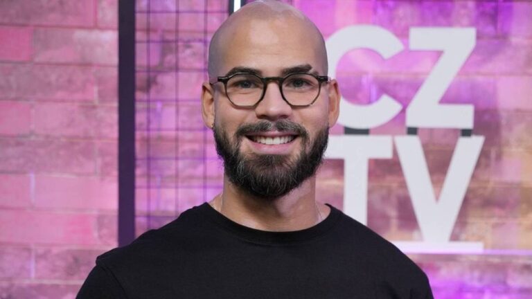 Homem careca de barba e óculos sorri diante de painel colorido com logotipo CZ TV em estúdio de televisão