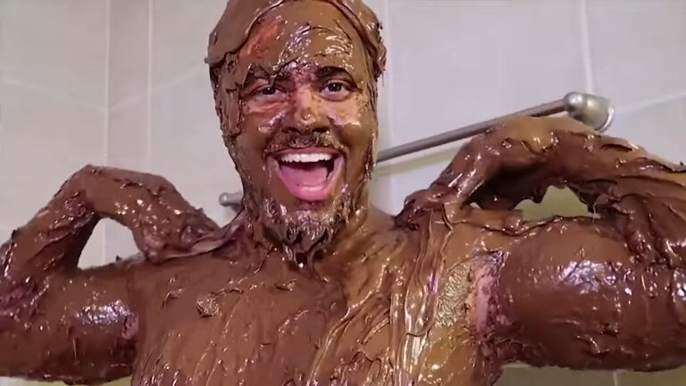 Luccas Neto coberto de chocolate sorri com mãos nos ombros em banheiro com azulejos claros em cena de desafio para canal na web