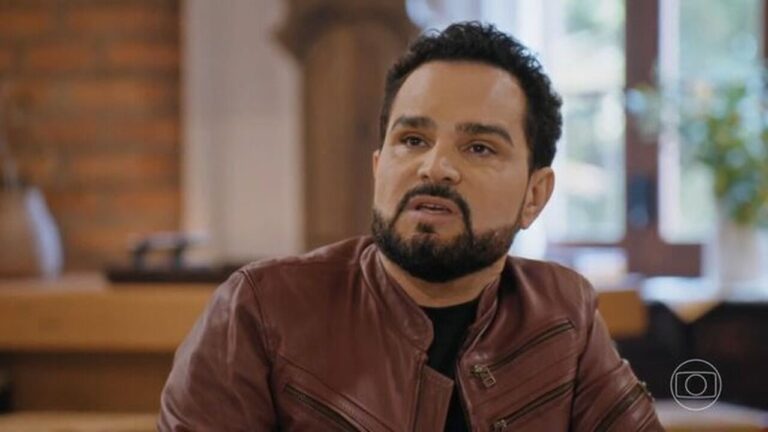 Homem de barba cheia e cabelo curto e escuro veste jaqueta de couro marrom e camiseta preta, com expressão séria, em ambiente interno iluminado por luz natural com parede de tijolos e janelas ao fundo.