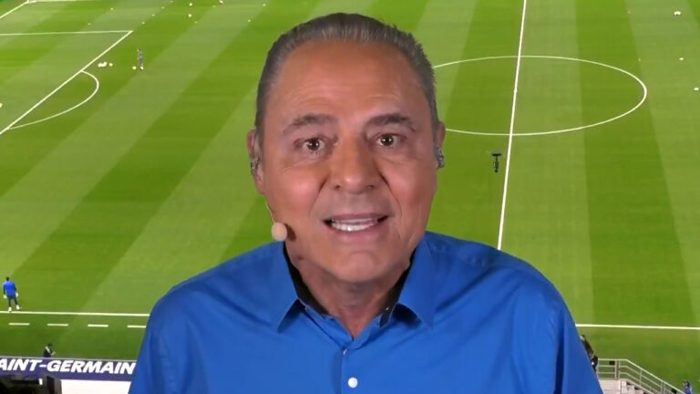 Luis Roberto sorri de camisa azul e headset com gramado de estádio ao fundo durante transmissão de jogo de futebol na Globo