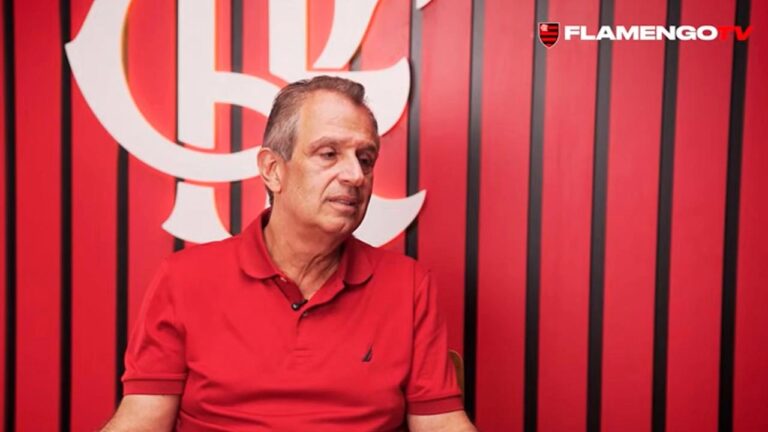 Homem adulto sentado em ambiente interno com fundo institucional do Flamengo, pele clara cabelos curtos grisalhos penteados para trás expressão séria e concentrada olhando levemente para o lado esquerdo da imagem, veste camisa polo vermelha de manga curta com pequeno logotipo no peito, segura as mãos próximas ao corpo em posição de conversa ou entrevista, atrás dele parede vermelha com listras verticais pretas e grande símbolo estilizado do Flamengo em branco parcialmente visível, no canto superior direito aparece a marca Flamengo TV, iluminação uniforme e frontal típica de estúdio ou sala de entrevistas transmite contexto de fala oficial ou depoimento relacionado ao clube