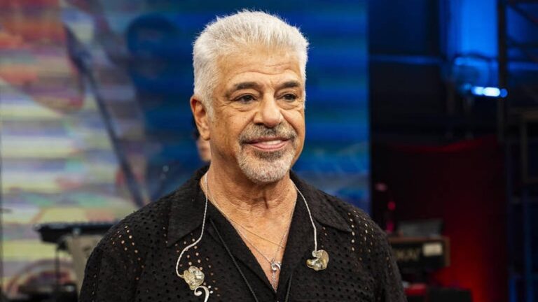 Homem de cabelos brancos curtos e barba grisalha sorri levemente enquanto usa camisa preta com textura furada e fones de ouvido de retorno presos por fios transparentes, em ambiente de estúdio com luzes coloridas e equipamentos musicais desfocados ao fundo