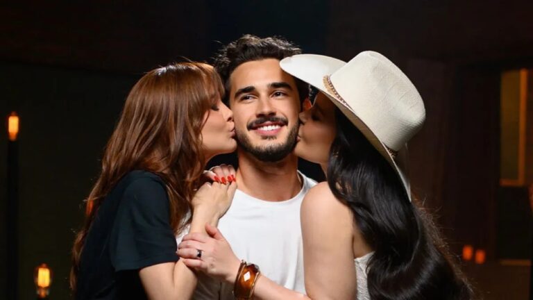 Três pessoas posam juntas em um ambiente interno iluminado. Um homem ao centro sorri enquanto duas mulheres, uma de cada lado, dão um beijo em suas bochechas. A mulher à esquerda tem cabelos castanhos e usa camiseta preta; a mulher à direita tem cabelos longos e escuros e usa um chapéu claro. Todos demonstram proximidade e alegria
