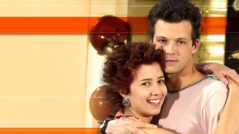 Um casal posa abraçado em ambiente interno decorado com balões. A mulher, de cabelo curto e avermelhado, sorri para a câmera, enquanto o homem, de cabelo escuro e expressão neutra, envolve seus ombros com o braço. Ambos estão em primeiro plano, com fundo desfocado em tons quentes e faixas gráficas horizontais em laranja e bege.