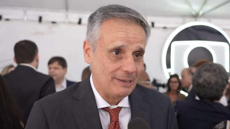 Manuel Belmar com terno escuro e gravata vermelha, falando a um microfone em evento, com o logo da Globo ao fundo