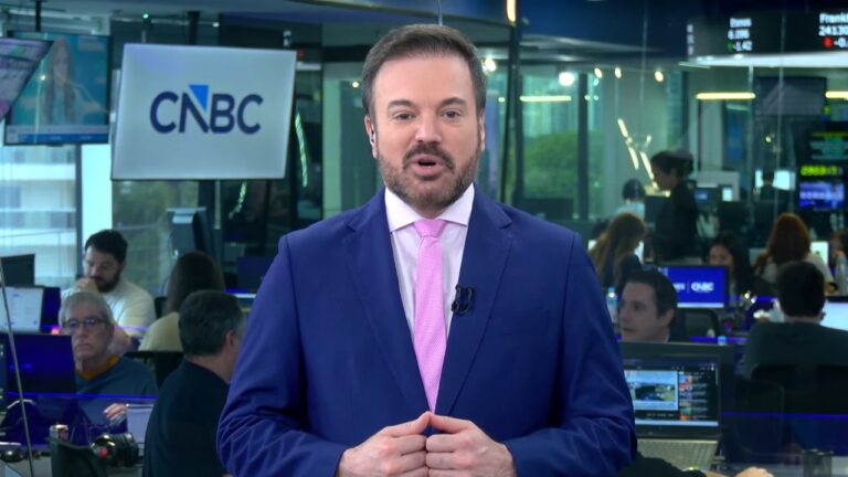 Apresentador de telejornal aparece em plano médio dentro de uma redação, usando terno azul e gravata rosa, enquanto fala para a câmera. Ao fundo, jornalistas trabalham em estações com computadores e telas exibem gráficos e informações do mercado. Um painel com o logotipo da CNBC está visível à esquerda, indicando um ambiente de notícias econômicas ao vivo.