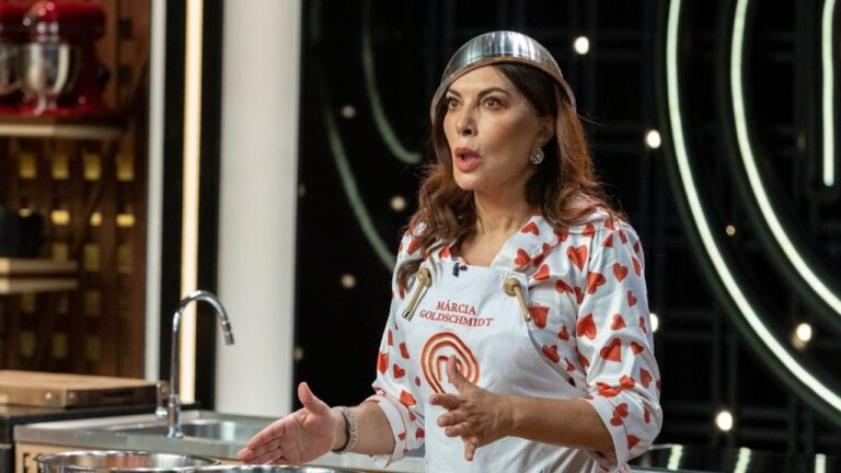 Participante de um programa culinário fala de forma expressiva enquanto gesticula. Ela veste avental branco com estampa de corações vermelhos e nome bordado, além de ter uma tigela de metal apoiada sobre a cabeça em tom humorístico. Ao fundo, vê-se a cozinha do estúdio com equipamentos e iluminação de cenário.