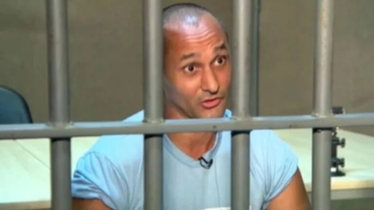 Homem de cabeça raspada e camiseta azul clara aparece atrás das grades de uma cela, olhando para frente com expressão séria, em ambiente interno iluminado com mesa e cadeira ao fundo