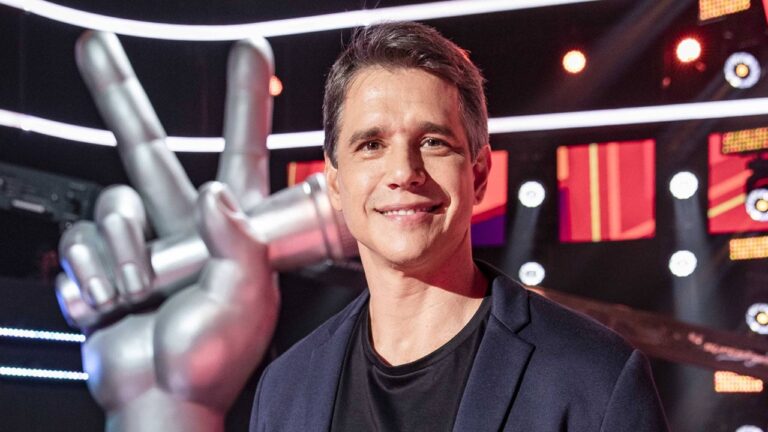 Homem sorridente posa em um estúdio iluminado, com luzes coloridas ao fundo e uma grande escultura prateada em forma de mão segurando um microfone. Ele usa camiseta preta e blazer escuro, olhando diretamente para a câmera