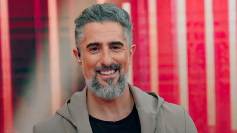 Homem de meia-idade sorrindo em um estúdio colorido, com barba grisalha bem aparada, cabelo curto também grisalho e usando jaqueta clara sobre camiseta escura. O fundo apresenta painéis verticais em tons vibrantes de vermelho e rosa.