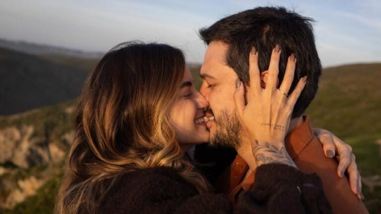 Casal abraçado ao ar livre, em meio a uma paisagem de colinas ao pôr do sol, trocando um beijo com os rostos encostados e olhos fechados. A mulher segura o rosto do homem com as duas mãos, exibindo tatuagens delicadas no pulso e anéis