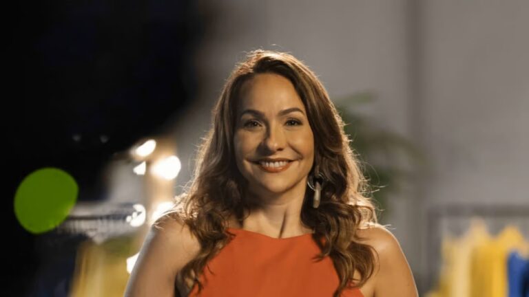 Mulher sorridente, com cabelos castanhos ondulados e vestido laranja, posa em ambiente interno iluminado; o fundo desfocado mostra luzes e elementos decorativos