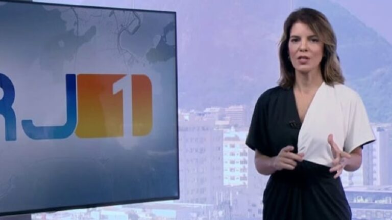 Uma apresentadora de telejornal aparece em estúdio, de pé, falando para a câmera. Ela usa um vestido bicolor em preto e branco e gesticula com as mãos. Ao seu lado, um telão exibe o logotipo “RJ1”. Ao fundo, através de uma janela, é possível ver prédios da cidade sob um céu claro, sugerindo um cenário urbano.
