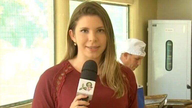 Repórter segura um microfone enquanto faz uma entrada ao vivo em um ambiente interno iluminado. Ela usa blusa vinho e está em primeiro plano, olhando para a câmera. Ao fundo, um profissional com touca branca trabalha próximo a uma bancada