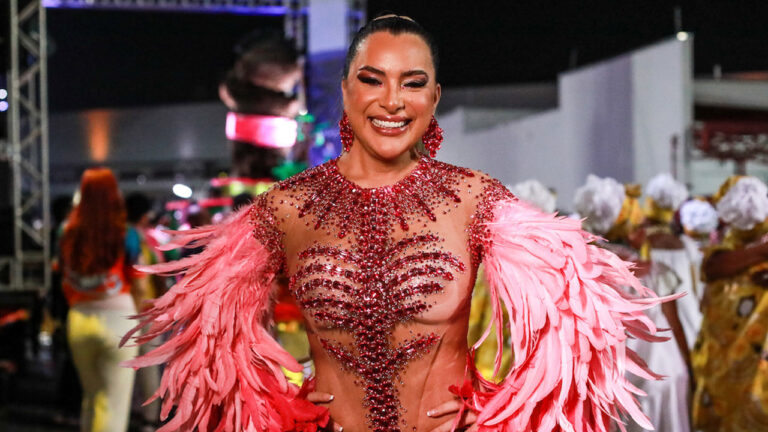 Marina Orlanda sorrindo com fantasia de carnaval com corpo transparente, pedrarias vermelhas e plumas rosas nos braços durante desfile
