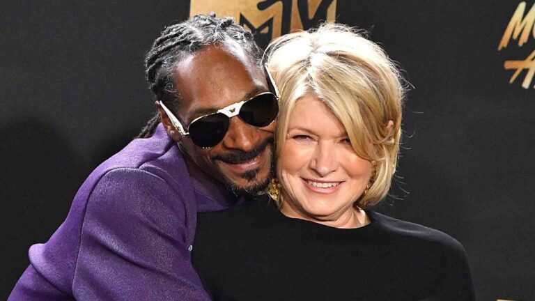 Homem de terno roxo e óculos escuros abraça mulher sorridente de roupa preta em evento da MTV com fundo preto e dourado