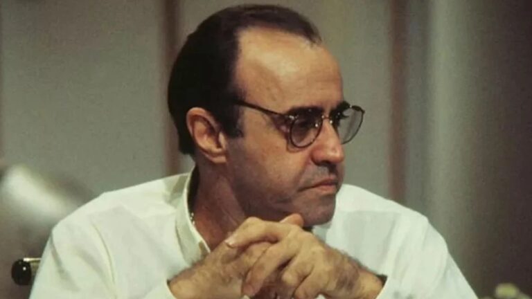 Homem de óculos e expressão séria, sentado com as mãos entrelaçadas, em ambiente interno com iluminação suave.