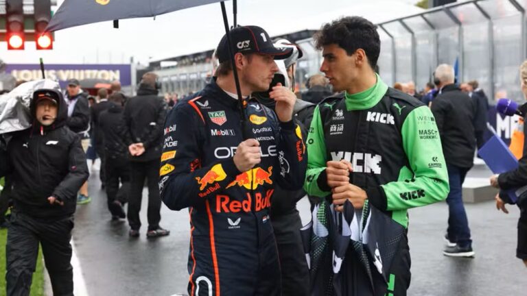 Dois pilotos de automobilismo conversam próximos a pista molhada, um vestindo macacão preto com logotipo da Red Bull e segurando guarda-chuva, o outro com macacão verde e preto, enquanto pessoas circulam ao fundo em ambiente de corrida
