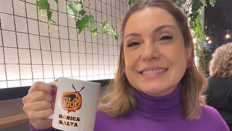 Monica Malta sorri segurando caneca com logo do Globo e seu nome em ambiente interno com plantas e iluminação aconchegante