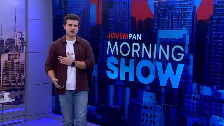 A imagem mostra André Marinho, um apresentador em pé no estúdio do programa Morning Show, com o logotipo exibido em um grande painel ao fundo. Ele veste camisa marrom aberta sobre camiseta branca e calça jeans, segurando um celular e usando um relógio inteligente no pulso. O cenário tem tons de azul e exibe prédios ao fundo, remetendo a uma paisagem urbana moderna
