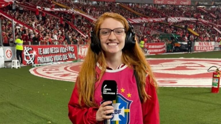 Jovem repórter ruiva, usando headphones grandes e colete vermelho de credenciamento, sorri enquanto segura um microfone em frente ao gramado de um estádio lotado. Faixas e bandeiras vermelhas ocupam as arquibancadas ao fundo, criando clima de partida de futebol