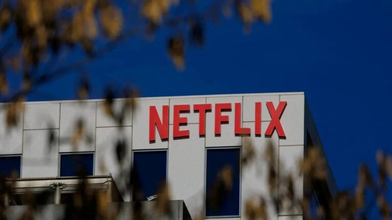Logotipo da Netflix em letras vermelhas na fachada de prédio corporativo sob céu azul com galhos desfocados em primeiro plano