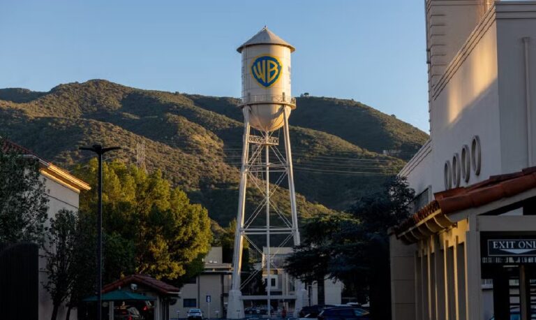 Caixa d'água metálica com o logotipo da Warner Bros. em destaque, erguida sobre uma torre alta no estúdio da empresa. Ao fundo, montanhas iluminadas pela luz do fim de tarde e prédios do complexo cinematográfico