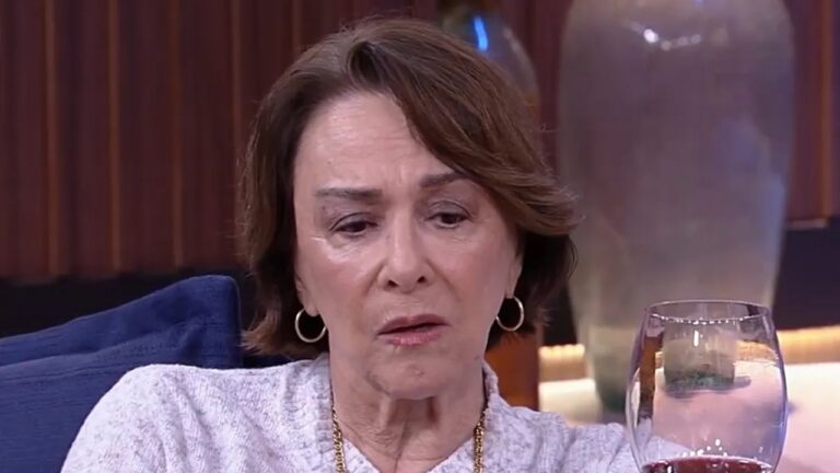 Mulher de meia-idade sentada em ambiente interno, com expressão séria e olhar baixo, segurando uma taça de vinho tinto. Ela usa brincos de argola e colar dourado, veste blusa clara e está apoiada em uma poltrona azul. Ao fundo, aparecem vasos decorativos e iluminação suave, sugerindo uma cena intimista, possivelmente de novela ou programa de televisão.