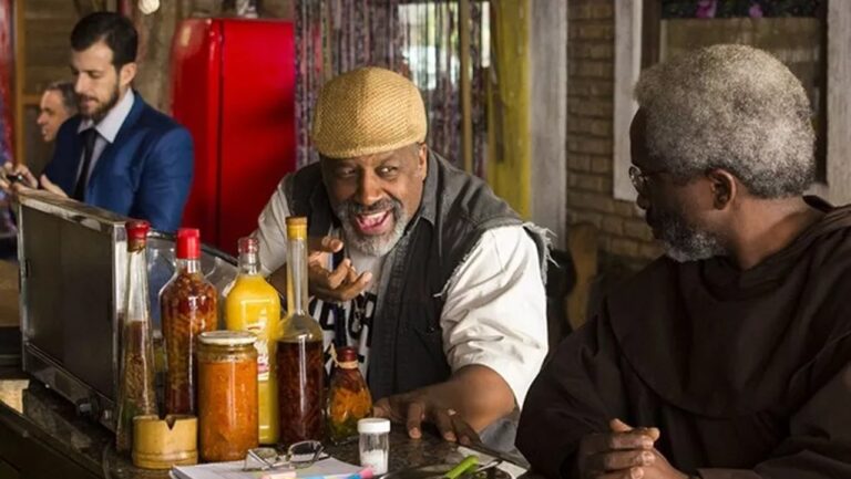 Dois homens conversam animadamente em um balcão com várias garrafas e potes à frente; um deles usa boina clara e sorri amplamente, enquanto o outro, de barba grisalha e roupas escuras, o observa com atenção.