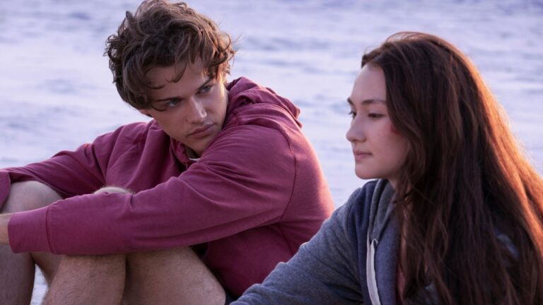 Dois jovens sentados lado a lado à beira da água, em um ambiente ao ar livre com o mar ao fundo. À esquerda, um jovem de cabelo curto e bagunçado veste moletom rosa e shorts, está com os joelhos dobrados e olha para a pessoa ao seu lado com expressão séria e reflexiva. À direita, uma jovem de cabelos longos e castanhos usa moletom cinza, olha para frente com semblante calmo e pensativo. A luz suave sugere fim de tarde, criando um clima íntimo e contemplativo.