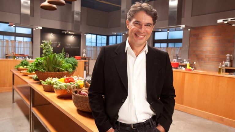 Homem sorridente em um estúdio de culinária, usando camisa branca e blazer preto, em pé diante de uma bancada de madeira com cestos de legumes e verduras frescas. O ambiente é amplo e bem iluminado, com cozinhas ao fundo, utensílios visíveis e aparência de programa gastronômico ou gravação televisiva.