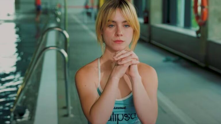 Mulher de maiô azul claro com mãos entrelaçadas em frente a piscina coberta iluminada por janelas grandes