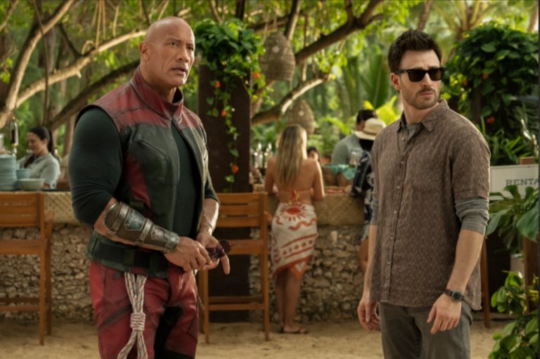 Dwayne Johnson e Chris Evans observam o ambiente em cena de filme ambientada em bar tropical ao ar livre com figurantes ao fundo