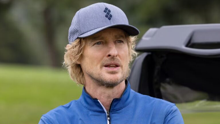 Owen Wilson usa boné e jaqueta azul em campo de golfe olhando atento com carrinho ao fundo em ambiente ao ar livre