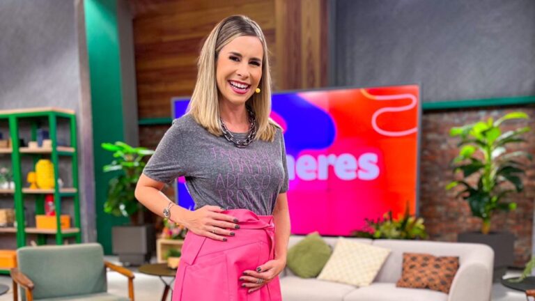 Apresentadora sorri em estúdio de TV usando camiseta cinza e saia rosa com cenário colorido e painel do programa ao fundo