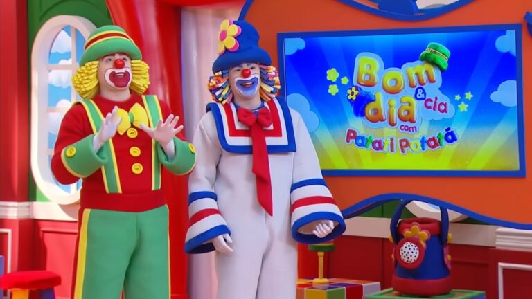 A imagem mostra os palhaços Patati e Patatá no cenário colorido do programa infantil Bom dia & Cia, exibido pelo SBT. Eles estão sorrindo e vestidos com seus trajes característicos — um de vermelho, verde e amarelo, e o outro de azul, branco e vermelho. Ao fundo, há uma televisão com o logotipo do programa sobre um fundo azul e amarelo com nuvens e estrelas, reforçando o ambiente alegre e lúdico