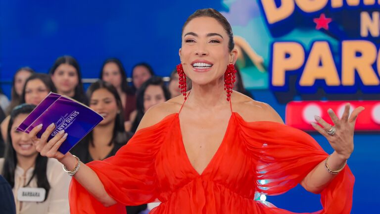 A imagem mostra Patricia Abravanel, uma apresentadora sorridente em um estúdio de TV colorido, com plateia ao fundo. Ela veste um vestido vermelho vibrante de mangas bufantes e usa brincos grandes da mesma cor. Em suas mãos, segura cartões roxos com o nome Silvio Santos. Atrás dela, há um painel com o título Domingo no Parque em letras grandes e coloridas