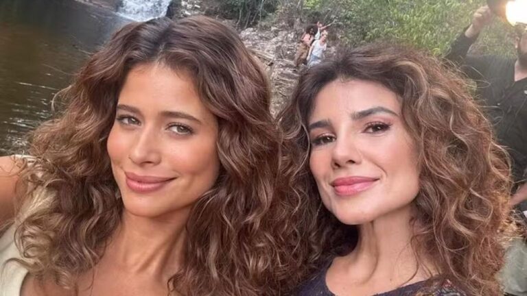Duas mulheres posam sorrindo para uma selfie em ambiente externo, com cabelos cacheados soltos e maquiagem leve; ao fundo, vê-se uma área de natureza com rio, pedras e uma pequena queda-d’água, além de parte de uma equipe de gravação desfocada.