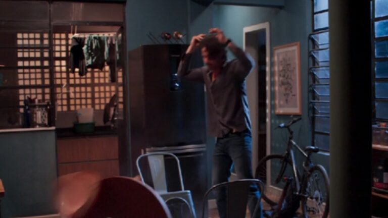 A imagem mostra um homem em um ambiente doméstico, aparentemente em um momento de desespero ou raiva. Ele está de pé, com as mãos na cabeça, e parece se mover de forma agitada. O cenário é uma casa ou apartamento simples, com cozinha ao fundo, cadeiras de metal e uma bicicleta encostada na parede. A iluminação é suave, com tons azulados, sugerindo uma cena dramática.